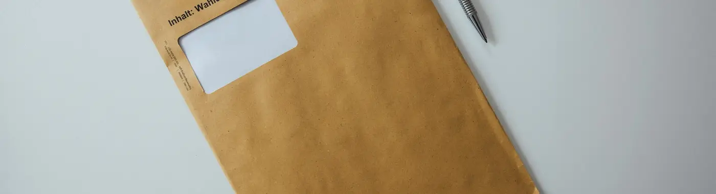 Ein brauner Briefumschlag mit einem Kugelschreiber liegt auf einem hellen, aufgeräumten Schreibtisch.