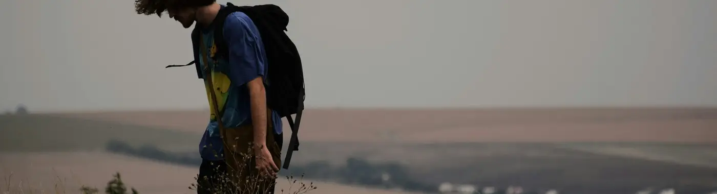 Eine Person mit Rucksack geht über einen offenen, grasbewachsenen Hügel mit weitem Blick am Horizont.